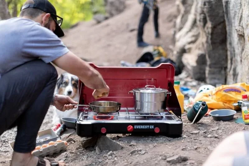 A man cooking food on a stove with dog for glamping experience.