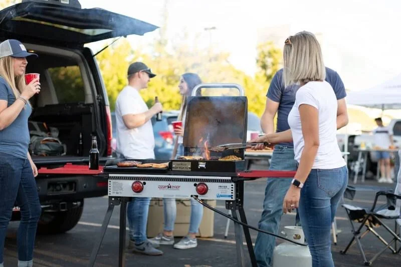 A group of people cooking food on a grill while tailgating with the Camp Chef Pro 14